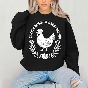 Può includere: Felpa nera con una grafica bianca di una gallina e il testo "Chicken Raising & Jesus Praising".