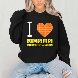 Puede incluir: Sudadera negra con el texto "I" y un coraz&oacute;n de gofre. Debajo del coraz&oacute;n, el texto "JESUS" se muestra en letras amarillas, con "JESUS YOU" debajo.