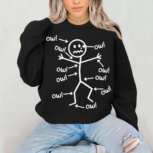 May include: Black sweatshirt with a white stick figure design. The stick figure has arrows pointing to it with the word "ow!" next to each arrow. The sweatshirt is a crew neck style.