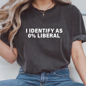 以下が含まれることがあります： 「I IDENTIFY AS 0% LIBERAL」の白文字がプリントされた濃いグレーのTシャツ。クルーネックのシャツは柔らかい素材で作られているようです。モデルは青いジーンズを着用しています。