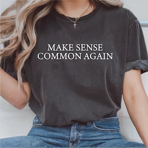 以下が含まれることがあります： 「MAKE SENSE COMMON AGAIN」の白文字がプリントされた濃いグレーのTシャツ。少し色あせた外観で、クルーネックです。袖は折り返されています。モデルはブルージーンズを着用しています。