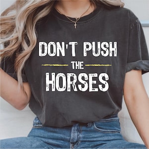 Puede incluir: Camiseta gris oscuro con el texto "DON'T PUSH THE HORSES" en letras blancas desgastadas. Una línea amarilla subraya "THE". La camiseta se lleva con vaqueros azules.