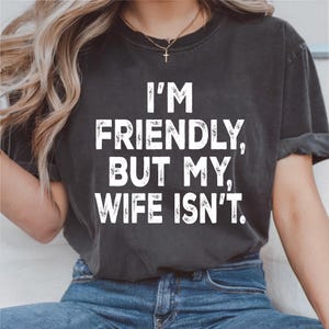 May include: Dark gray t-shirt with the text "I'M FRIENDLY, BUT MY, WIFE ISN'T." in white block letters. The shirt is a crew neck style and has short sleeves. The model is wearing blue jeans.