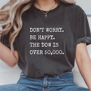 May include: Dark gray t-shirt with the text "DON'T WORRY. BE HAPPY. THE DOW IS OVER 50,000." in white block letters. The shirt is worn with blue jeans.
