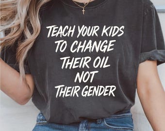 Op de afbeelding: Donkergrijs t-shirt met de witte tekst "TEACH YOUR KIDS TO CHANGE THEIR OIL NOT THEIR GENDER." Het shirt wordt gedragen met een blauwe spijkerbroek. Een gouden ketting met een kruisvormige hanger is zichtbaar.