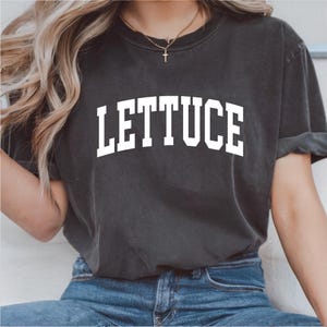 May include: Dark gray t-shirt with the word "LETTUCE" printed in large, white, block letters. The shirt has short sleeves and a crew neck. The model is wearing blue jeans.