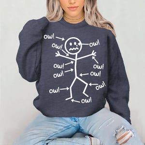 Puede incluir: Sudadera azul marino con un dise&ntilde;o de figura de palo blanca. La figura tiene flechas que apuntan a diferentes partes del cuerpo, cada una con la palabra "ow!". Un dise&ntilde;o humor&iacute;stico y casual.