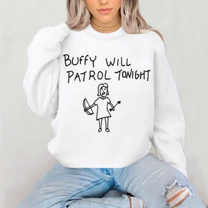 May include: White sweatshirt featuring the phrase "Buffy Will Patrol Tonight" in black, handwritten text. Beneath the text is a black and white illustration of a person holding a bow and arrow. The person is wearing a dress.
