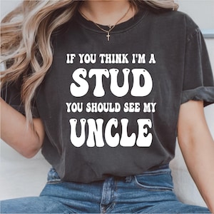 Pode incluir: T-shirt cinzenta escura com o texto branco "IF YOU THINK I'M A STUD YOU SHOULD SEE MY UNCLE". A camisa tem gola redonda e mangas curtas. O texto está em negrito, com um estilo retro.