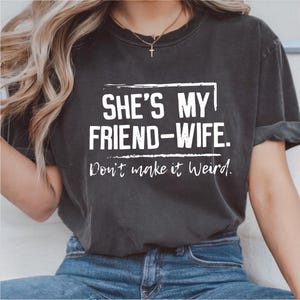 Puede incluir: Camiseta gris oscuro con el texto blanco "SHE'S MY FRIEND-WIFE. Don't make it Weird." en una fuente desgastada. La camiseta es de estilo casual, adecuada para el uso diario.
