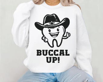Op de afbeelding: Wit sweatshirt met een cartoon tand die een cowboyhoed draagt en een duim omhoog geeft. De tekst "BUCCAL UP!" staat eronder in vette, zwarte letters. Het sweatshirt is een casual, comfortabele stijl.