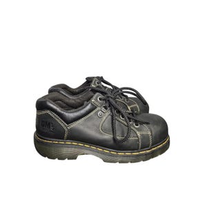 May include: Black leather work shoes with black laces and yellow stitching. The shoes have a thick sole and the brand name "DM's" is visible on the side.