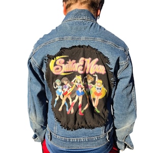 Könnte beinhalten: Blaue Jeansjacke mit einem großen schwarzen Aufnäher auf dem Rücken mit den Worten "Sailor Moon" in Gelb und Rosa sowie Illustrationen der Sailor Moon-Charaktere.