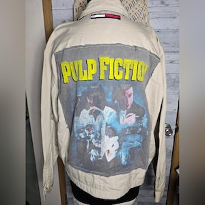 May include: Tan jacket with a graphic of the movie "Pulp Fiction" on the back. The graphic features the movie title in yellow letters and a scene from the film. The jacket has a collar and a Tommy Hilfiger logo.