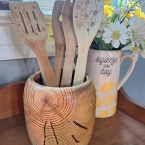 Peut inclure: Un pot à ustensiles en bois avec un fini grain de bois naturel, contenant plusieurs ustensiles de cuisine en bois. Les ustensiles comprennent une spatule fendue, une fourchette et une cuillère avec un motif floral. Une tasse en céramique avec le texte "squeeze the day" et un arrangement floral sont visibles.