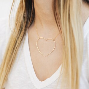 May include: A gold-colored heart-shaped pendant necklace. The pendant is a simple outline of a heart, suspended from a delicate chain. The necklace is worn against a white v-neck shirt.