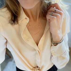 May include: A gold-toned heart-shaped pendant necklace. The necklace is worn with a cream-colored button-down shirt and multiple rings and bracelets. The shirt is tied at the waist. The jewelry is a simple, elegant style.