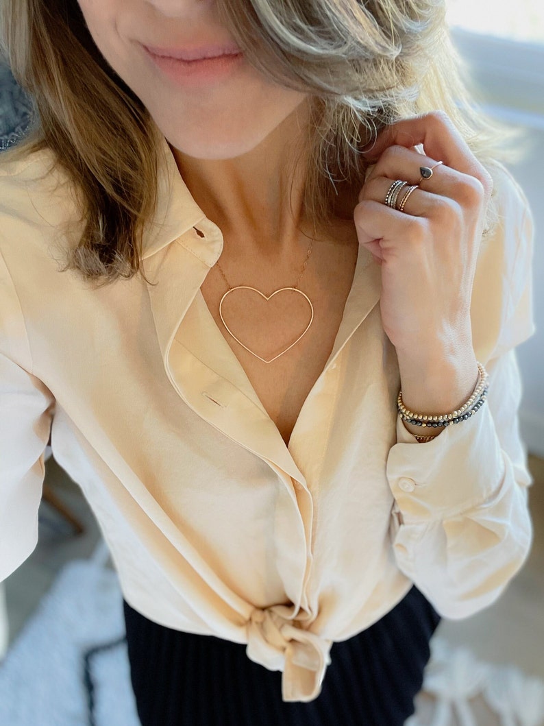 May include: A gold-toned heart-shaped pendant necklace. The necklace is worn with a cream-colored button-down shirt and multiple rings and bracelets. The shirt is tied at the waist. The jewelry is a simple, elegant style.