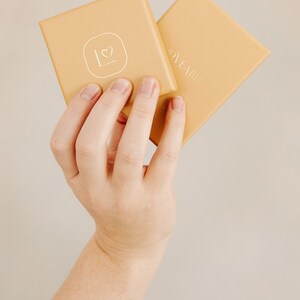 May include: Two square, light-orange boxes are held up. The top box has a white heart and the letter "L" inside a square. The other box has the word "LOVEABLE" in silver lettering. The boxes are held by a person.