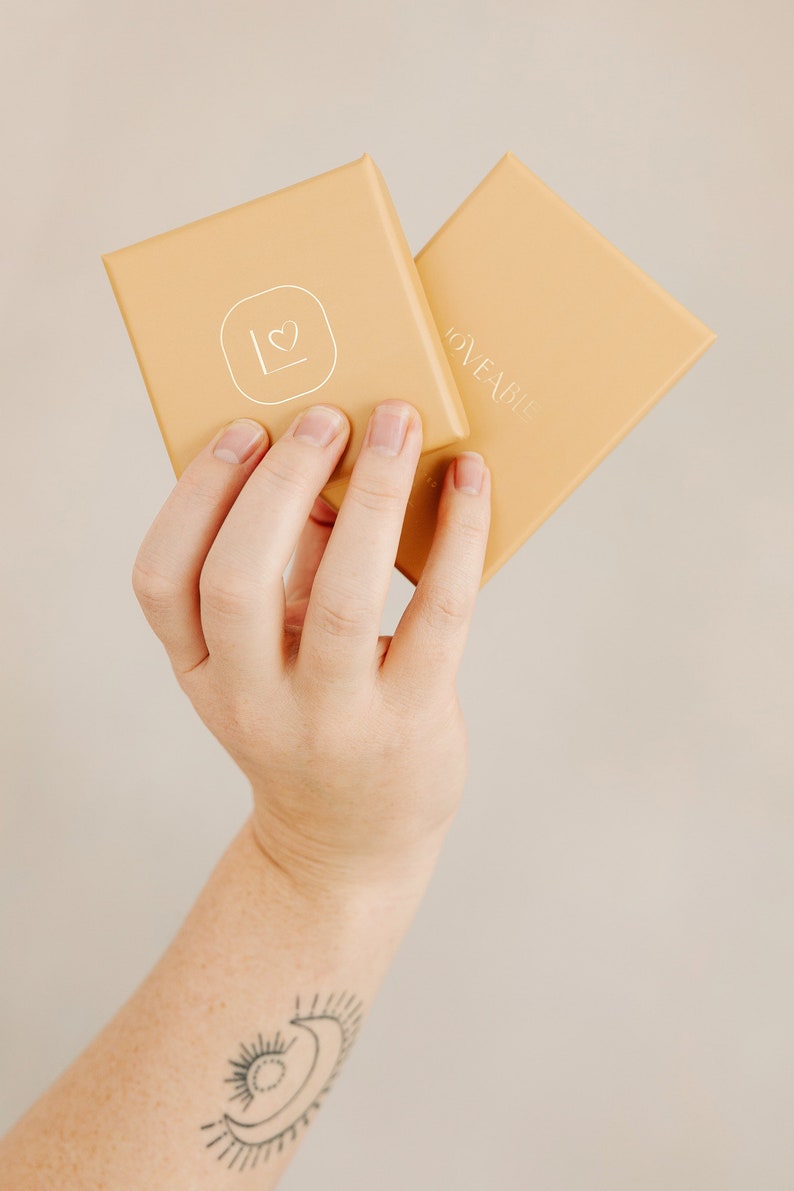 May include: Two square, light-orange boxes are held up. The top box has a white heart and the letter "L" inside a square. The other box has the word "LOVEABLE" in silver lettering. The boxes are held by a person.