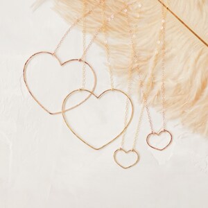 May include: Four gold-colored heart-shaped necklaces of varying sizes hang against a white background. The necklaces are suspended from delicate chains. A feathery, light brown accent is in the background.