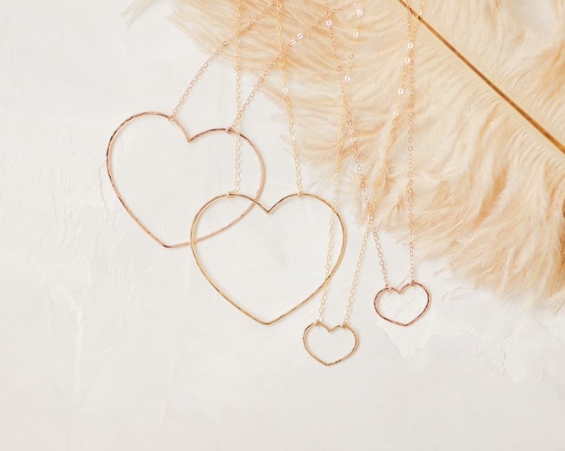 May include: Four gold-colored heart-shaped necklaces of varying sizes hang against a white background. The necklaces are suspended from delicate chains. A feathery, light brown accent is in the background.