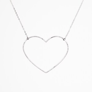 May include: A silver-colored necklace featuring a large, open heart pendant. The heart is crafted from a thin, continuous line, suspended from a delicate chain. The design is minimalist and modern, set against a plain white background.