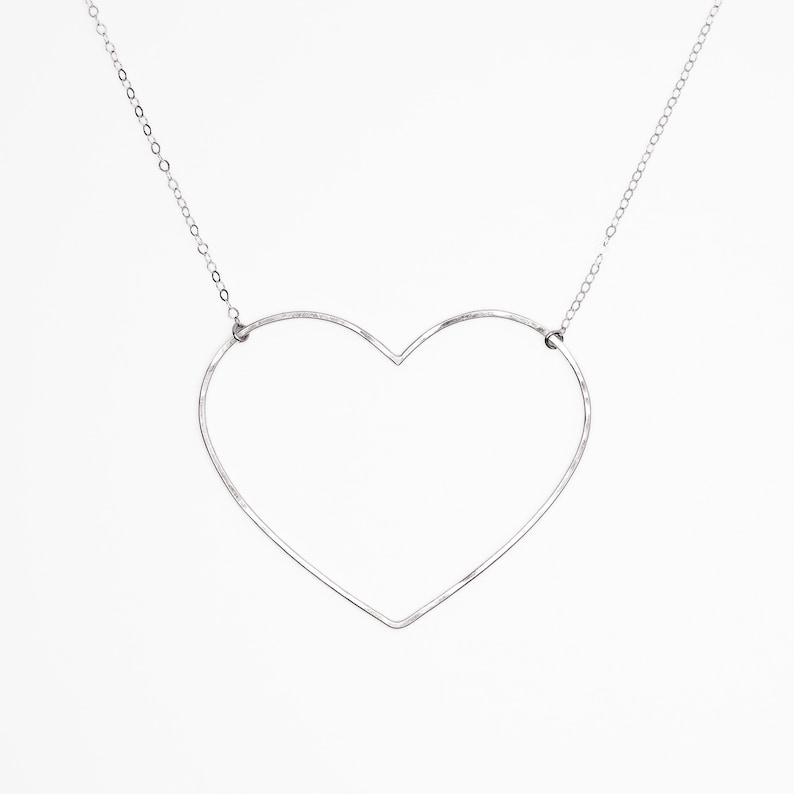 May include: A silver-colored necklace featuring a large, open heart pendant. The heart is crafted from a thin, continuous line, suspended from a delicate chain. The design is minimalist and modern, set against a plain white background.