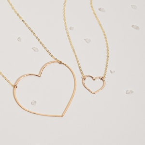 May include: Two gold-colored necklaces with heart-shaped pendants. The larger heart pendant is open, while the smaller one is solid. The necklaces are on a white surface with scattered, small, clear stones. The chains are delicate.