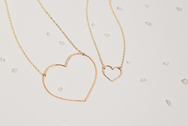May include: Two gold-colored necklaces with heart-shaped pendants. The larger heart pendant is open, while the smaller one is solid. The necklaces are on a white surface with scattered, small, clear stones. The chains are delicate.