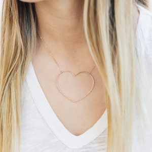 May include: A rose gold heart-shaped pendant necklace. The open heart design hangs from a delicate chain. The necklace is worn over a white V-neck shirt. The model has long blonde hair.