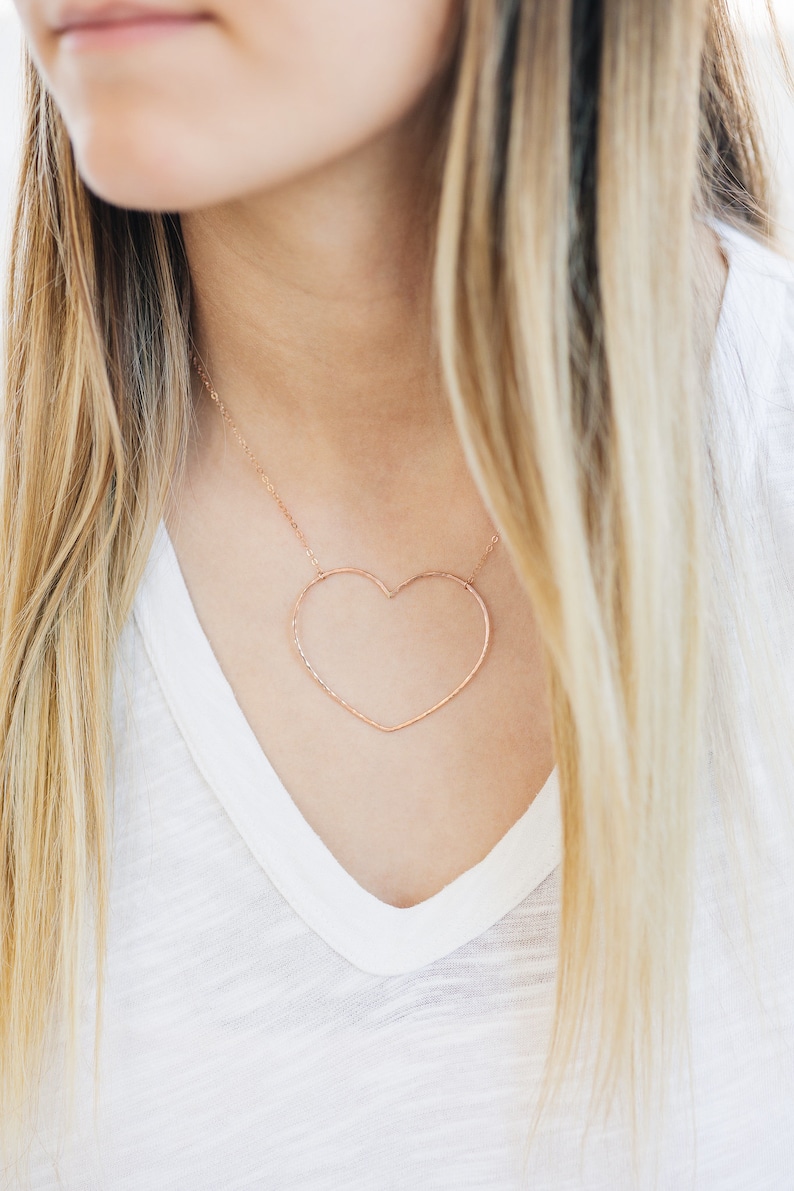 May include: A rose gold heart-shaped pendant necklace. The open heart design hangs from a delicate chain. The necklace is worn over a white V-neck shirt. The model has long blonde hair.