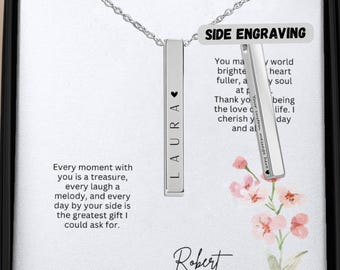 May include: A silver necklace with two vertical bars. The bar on the left has the name "LAURA" engraved on it. The bar on the right has the text "You make my world brighter, my heart fuller, at peace. Thank you for the love you cherish and always share." engraved on it. The necklace is displayed in a black box with a white background and a floral design.