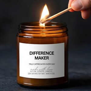 May include: A lit soy wax candle in a brown glass jar. The candle has a white label with the text "DIFFERENCE MAKER" and "TRULY APPRECIATED EVERY DAY". A match is held near the wick, igniting the flame. The candle is made in the USA.
