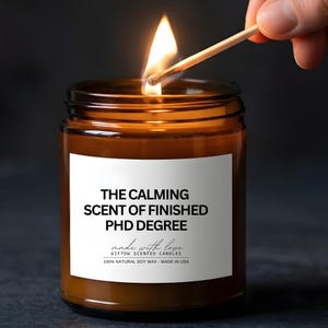 May include: A lit brown glass jar candle with a white label. The label reads "THE CALMING SCENT OF FINISHED PHD DEGREE". A match is held near the wick, igniting the flame. The candle is made with 100% natural soy wax.