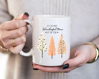 Puede incluir: Taza de cerámica blanca con un diseño en blanco y negro de tres árboles estilizados. El texto "It's the most Wonderful time of the year" está impreso en la taza.