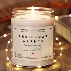 May include: A lit, cream-coloured scented candle in a glass jar. The label reads "CHRISTMAS WARMTH" and "made with love." The candle is 255g and has a burning time of 50-60 hours. A string of fairy lights encircles the candle, adding a warm glow.