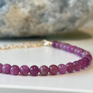 Può includere: Un delicato braccialetto con piccole pietre preziose rosa rotonde. Il braccialetto ha una catena e una chiusura color oro. Le pietre preziose sono di una vibrante tonalità di rosa e il braccialetto è esposto su una superficie bianca.