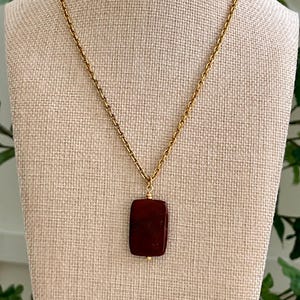 May include: A gold-colored chain necklace with a rectangular, dark red pendant. The pendant is a solid color and has rounded corners. The necklace is displayed on a beige fabric surface.