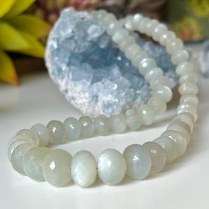 May include: A beaded necklace with pale green, faceted beads. The necklace is displayed on a white surface, with a blue crystal and green plants in the background. The beads have a subtle sheen.