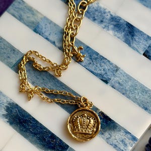 May include: Gold-toned necklace featuring a round pendant with a detailed crown design. The chain has a delicate link pattern and a fleur-de-lis charm. The necklace rests on a blue and white striped surface.