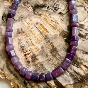 May include: A necklace composed of faceted, rectangular purple beads. The beads vary in shade from light lavender to deep violet. The necklace is displayed against a backdrop of a light brown, textured surface.