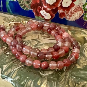 May include: A necklace made of round, reddish-pink beads with a gold clasp. The beads have a translucent quality, with variations in color from light pink to deep red. The necklace is displayed on a surface with a marbled pattern.