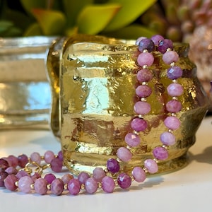 May include: A gold-colored, textured ceramic container with a handle is the backdrop for a necklace. The necklace features faceted, pink and purple gemstones strung together with gold-colored beads and clasps. The gemstones vary in shade.