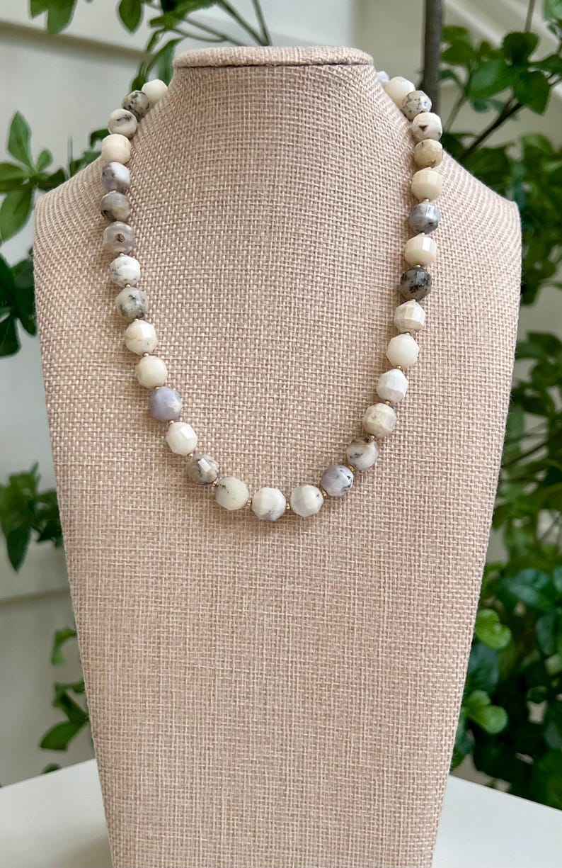 May include: A beaded necklace with a variety of neutral-toned beads, including white, gray, and beige. The beads are faceted and strung together, creating a textured look. The necklace is displayed on a beige jewelry bust.