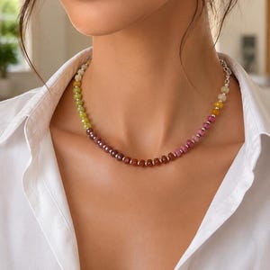 May include: A multi-colored gemstone necklace with a silver clasp. The necklace features a gradient of colors, including yellow, green, pink, and brown beads. The necklace is worn against a white collared shirt.
