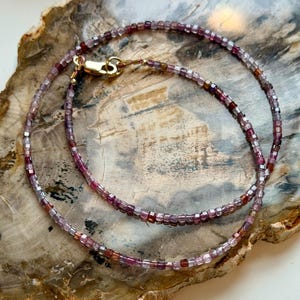 May include: A delicate necklace composed of small, square-cut, multi-colored beads in shades of pink, purple, and brown. The necklace is secured with a gold-colored clasp. The jewelry is displayed on a light brown, textured surface.