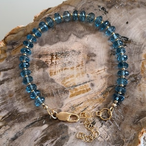 May include: A delicate bracelet featuring faceted, light blue gemstones strung together with gold-colored beads. The bracelet is secured with a gold-colored clasp and chain. The jewelry is displayed on a light brown surface.