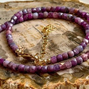 May include: A necklace featuring faceted, purplish-pink gemstones strung together. The necklace has a gold-colored clasp and chain. The gemstones are arranged in a continuous loop, creating a layered effect. The necklace is displayed on a textured, light brown surface.