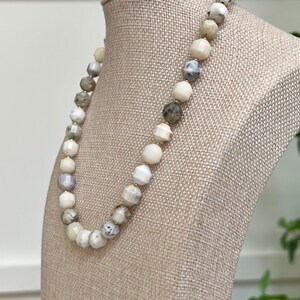 May include: A beaded necklace with a variety of neutral-toned beads, including white, gray, and cream colors. The beads are round and faceted, strung together with gold-colored spacers. The necklace is displayed on a beige mannequin.